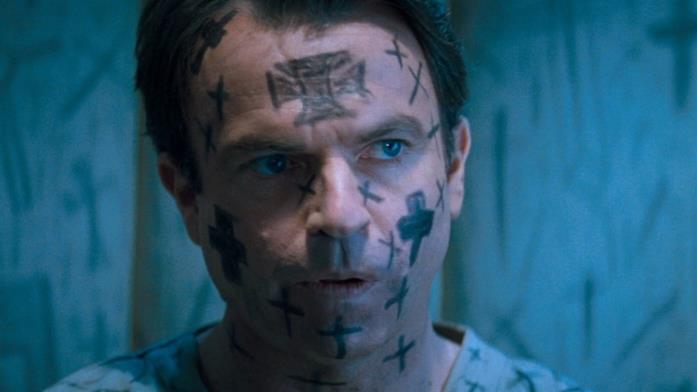 Il seme della follia, di John Carpenter (1994) John Trent (Sam Neill) con la faccia ricoperta di croci in un manicomio