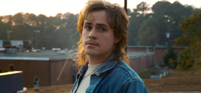 Billy Hargrove al suo primo giorno di scuola in Stranger Things 2 Billy Hargrove con giubbino di jeans e maglietta grigia, con la scuola di Hawkins sullo sfondo