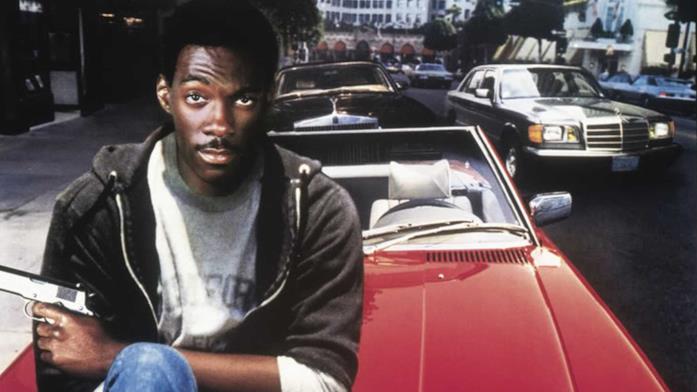 Eddie Murphy nei panni di Axel Foley con una pistola in mano seduto sul cofano di una auto rossa
