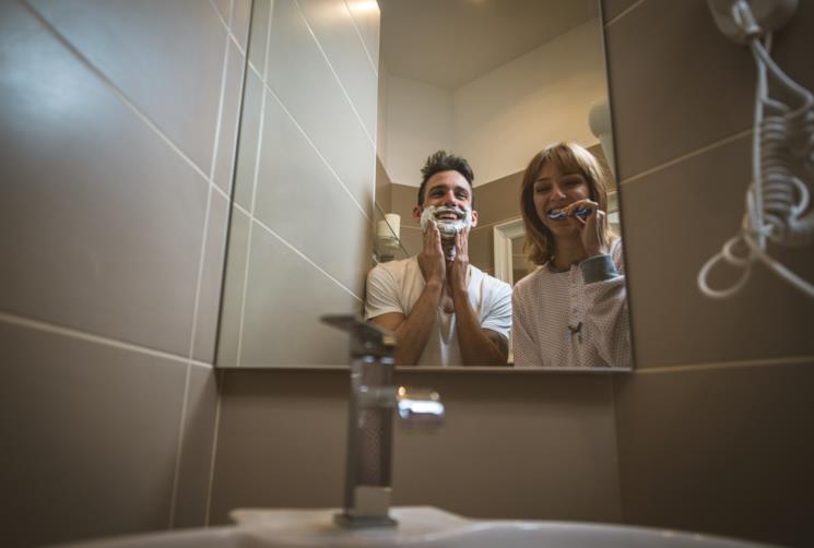 Coppia in bagno riflessa dallo specchio contenitore. Lui si fa la barba, lei si lava i denti